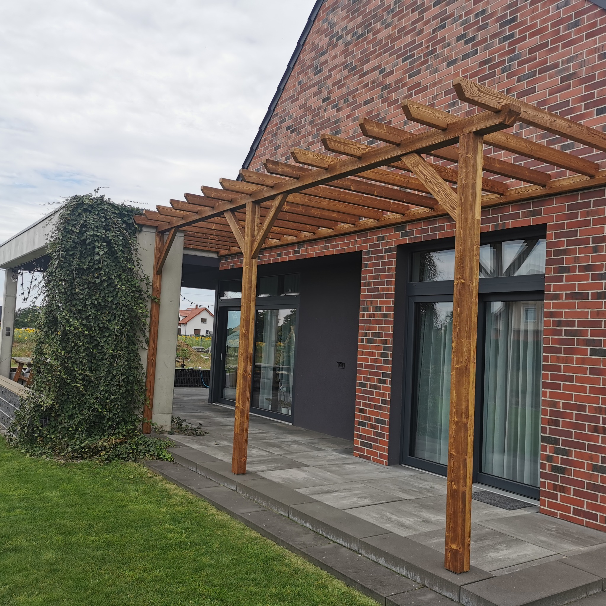 Drewniana pergola z belek przy ceglanym budynku. Na pierwszym planie trawnik, w tle budynek z oknami i kolumna porośnięta bluszczem. Całość w naturalnym oświetleniu.