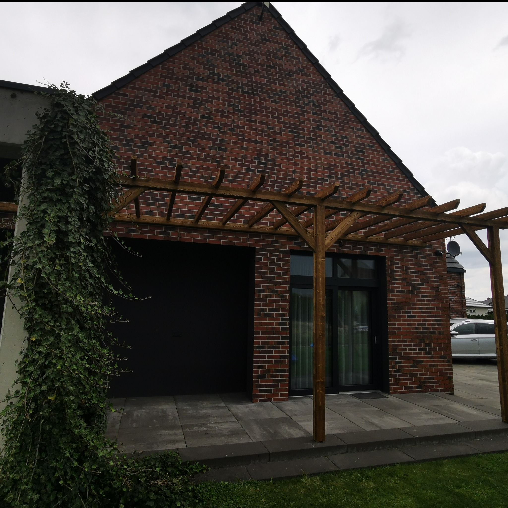 Drewniana pergola z elementami ozdobnymi, zlokalizowana przy ceglanym domu w Brzegu Dolnym. Konstrukcja częściowo porośnięta bluszczem, widoczne szare płyty tarasowe.