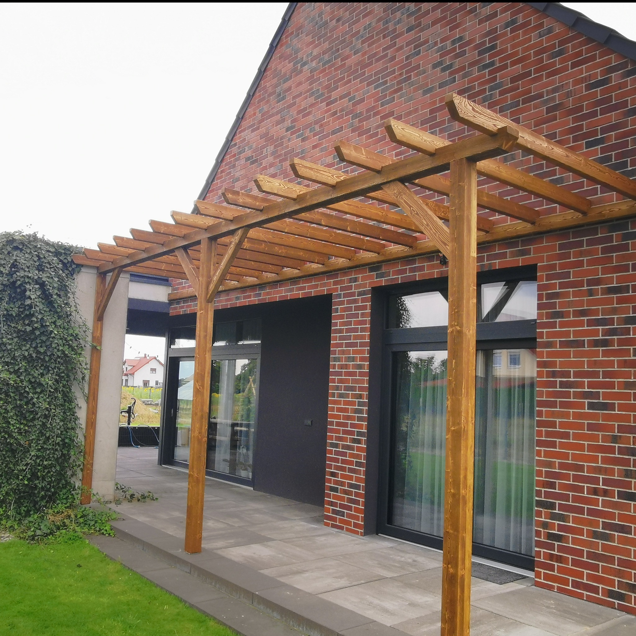 Drewniana pergola przy nowoczesnym domu z cegły. Konstrukcja z belek, widoczne słoje drewna. Szara kostka brukowa, fragment trawnika i zieleni. Dwa okna.