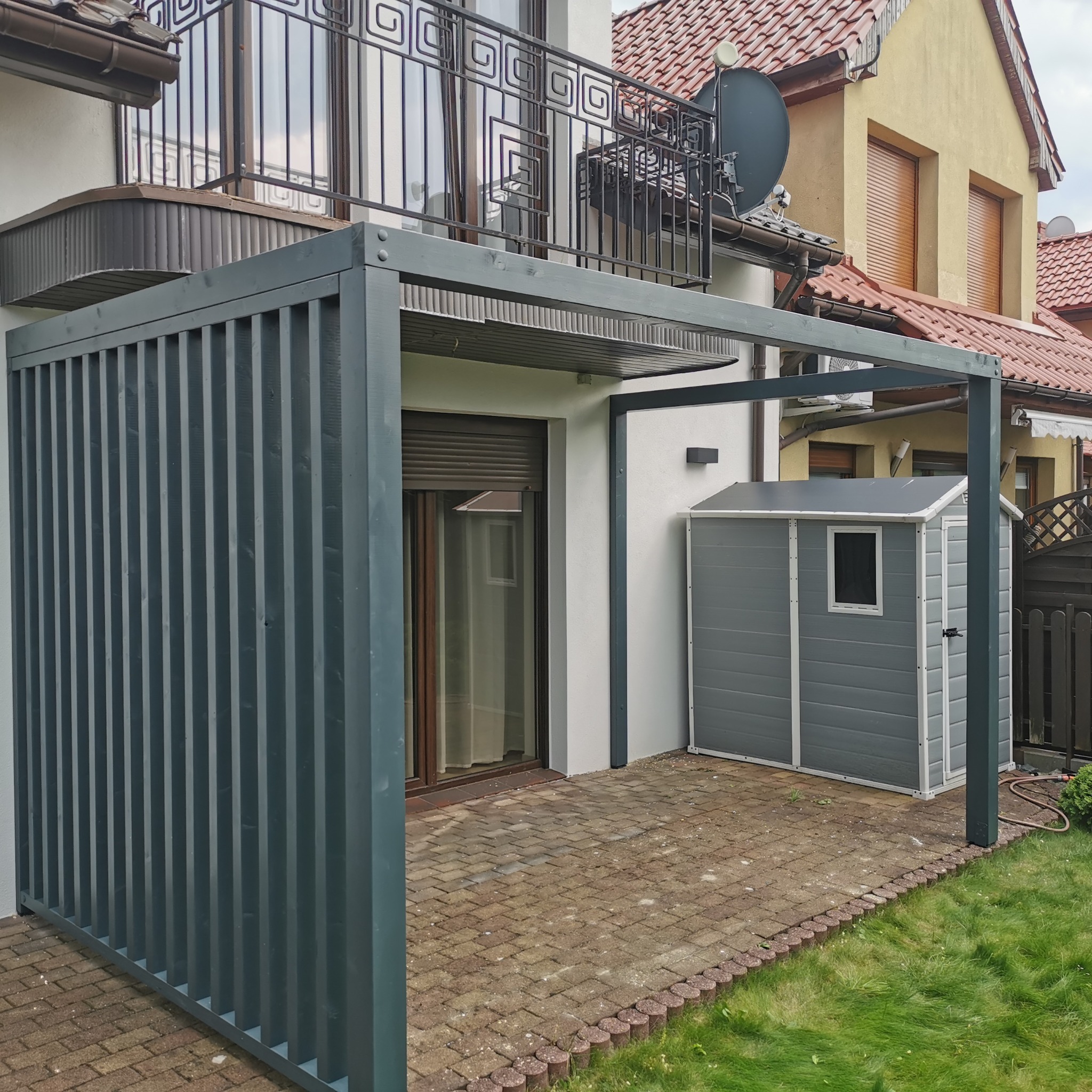 Szara pergola z pionowymi listwami, przylegająca do domu z balkonem i szarą komórką narzędziową na kostce brukowej. Widok z poziomu trawnika.