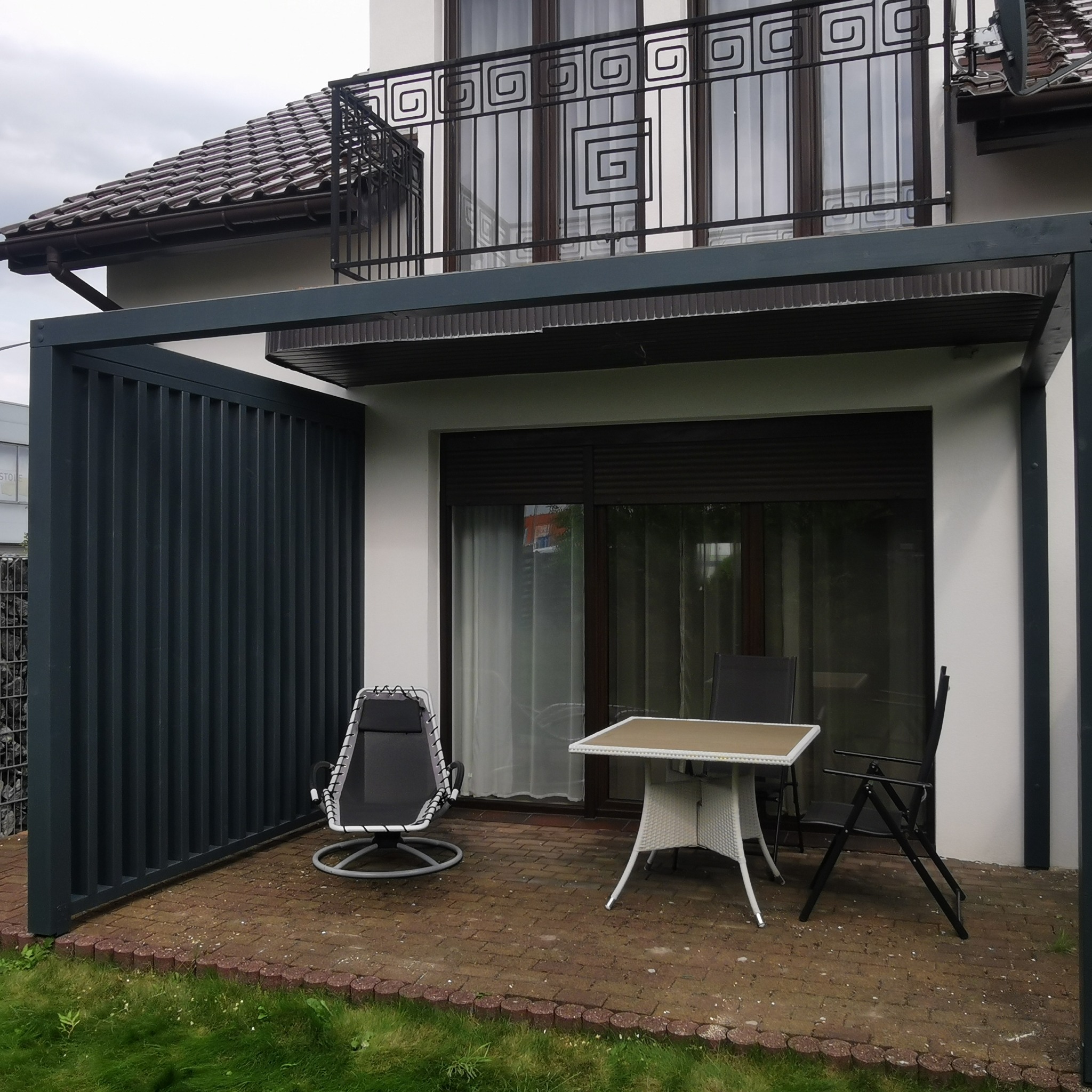 Nowoczesna, grafitowa pergola z metalu przy białym domu. Na tarasie stół i krzesła. Balustrada balkonu z geometrycznym wzorem.