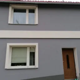 Szara elewacja dwukondygnacyjnego domu z białymi obramowaniami okien i poziomym pasem, brązowe drzwi wejściowe z długim, wąskim przeszkleniem, roleta w oknie na parterze.