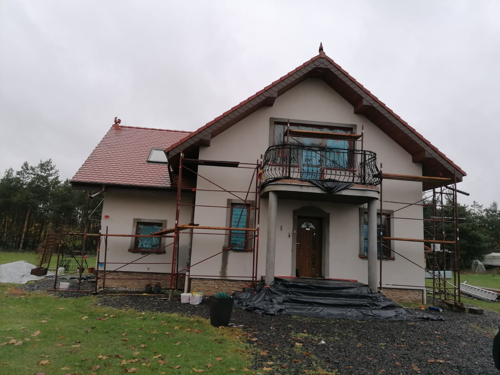 Dom jednorodzinny w trakcie prac elewacyjnych, widoczne rusztowania, okna zabezpieczone folią, częściowo ocieplona i pomalowana fasada, dach z czerwonej dachówki, figurka koguta na szczycie dachu.
