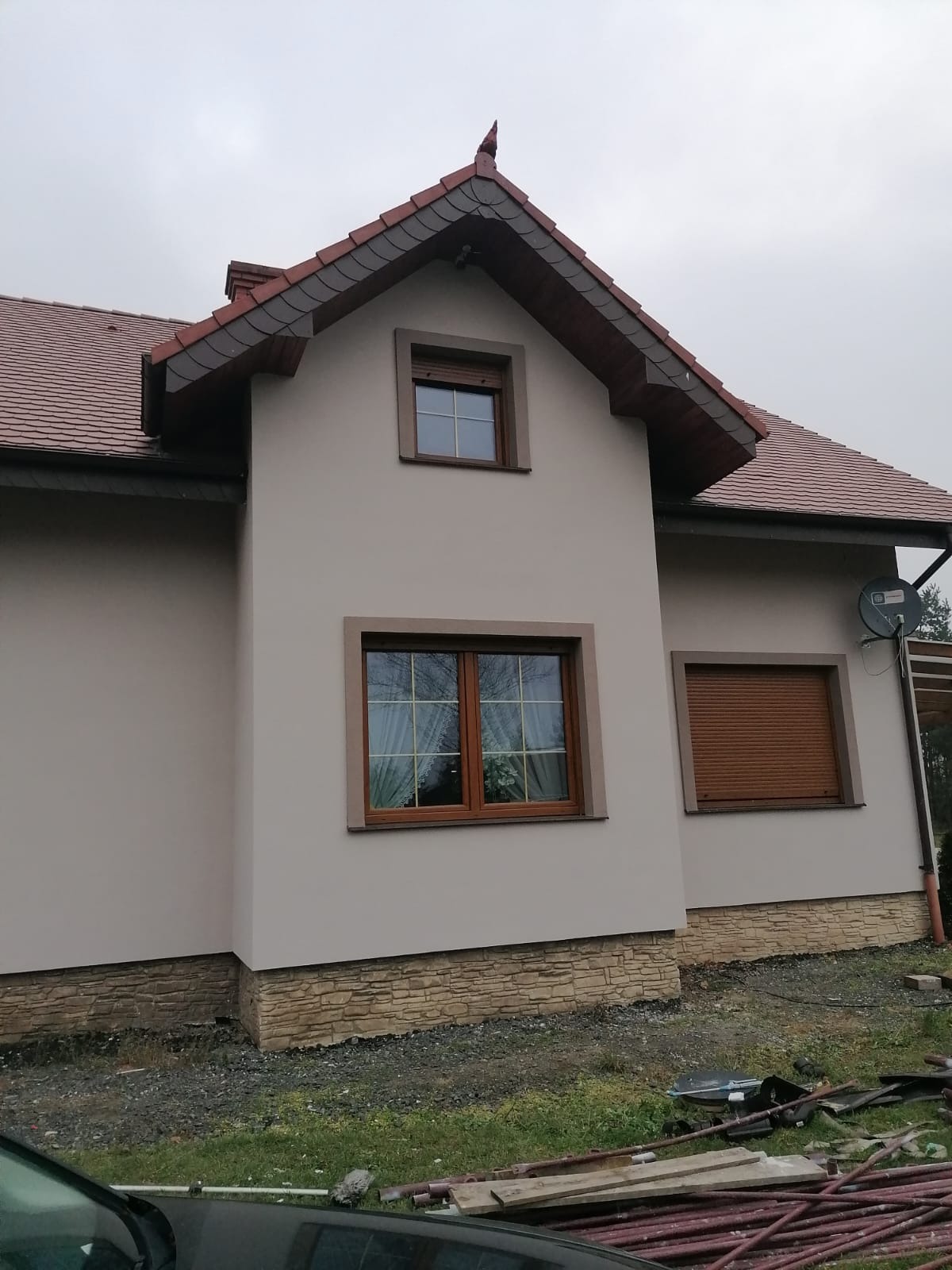 Elewacja domu jednorodzinnego z beżowym tynkiem, drewnianymi oknami z brązowymi ramami i roletami zewnętrznymi, wykończona kamieniem dekoracyjnym na dole, widoczna antena satelitarna na ścianie...