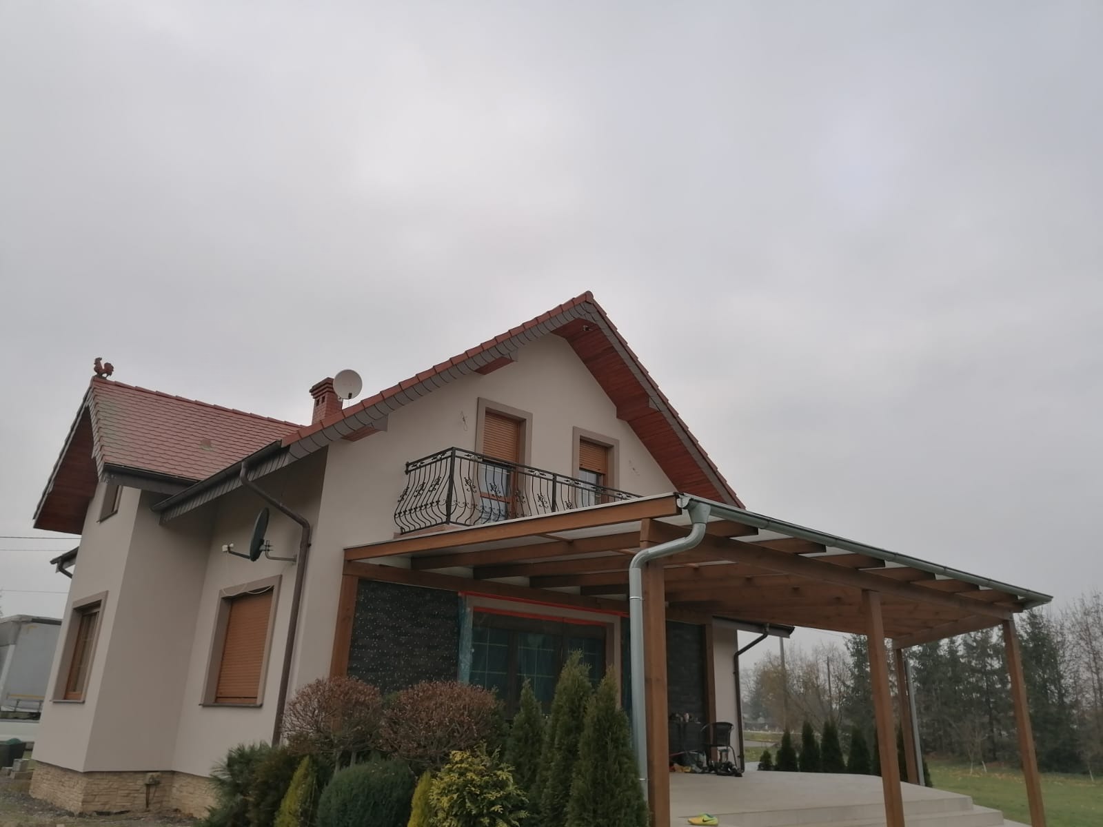 Dom jednorodzinny z jasną elewacją, czerwonym dachem, balkonem z ozdobną balustradą i drewnianą pergolą przy wejściu, sfotografowany w pochmurny dzień.