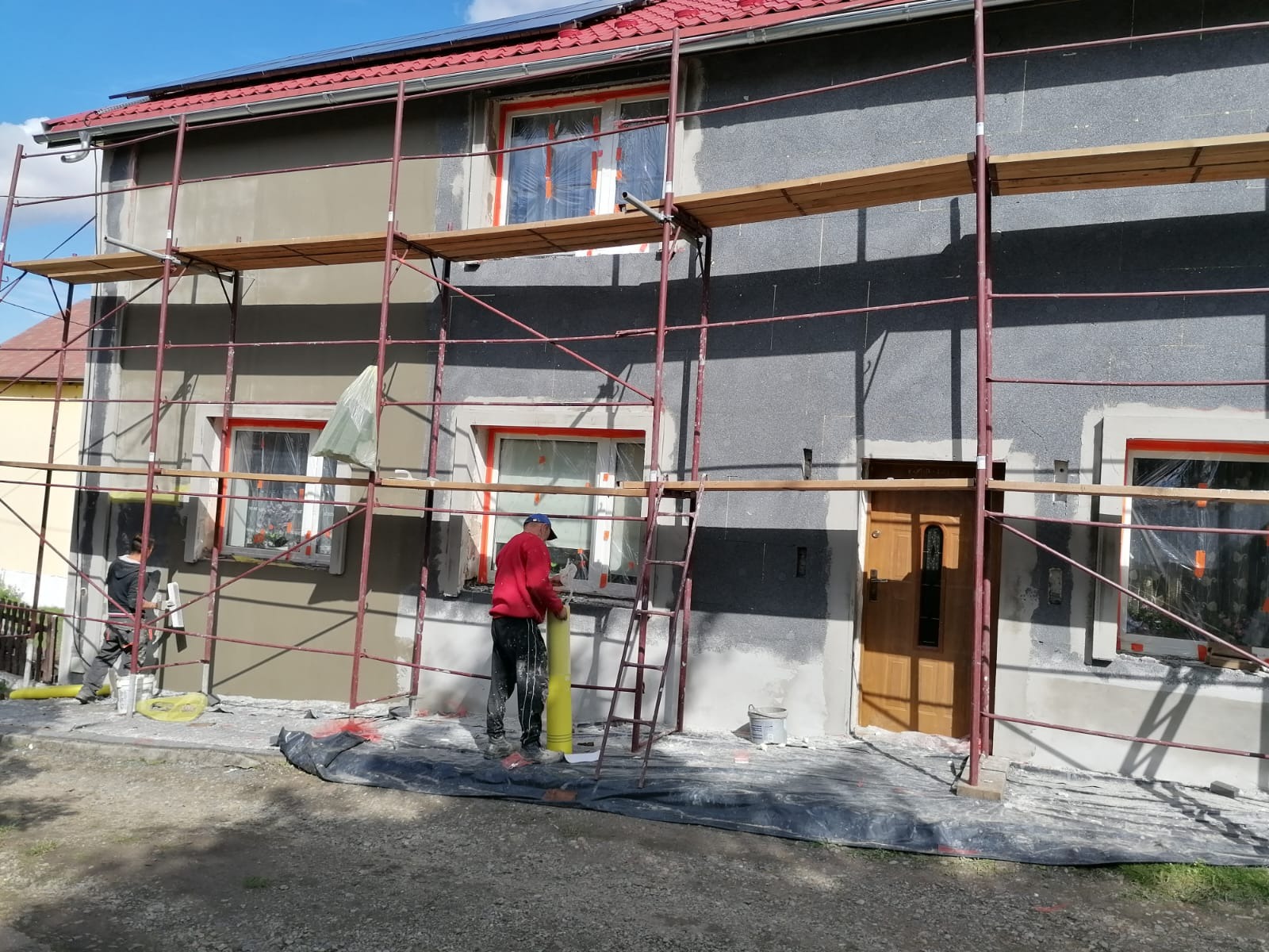 Ocieplanie elewacji domu jednorodzinnego z użyciem rusztowania, widoczne płyty styropianowe i częściowo nałożony klej, pracownik w czerwonej bluzie przygotowuje materiał.