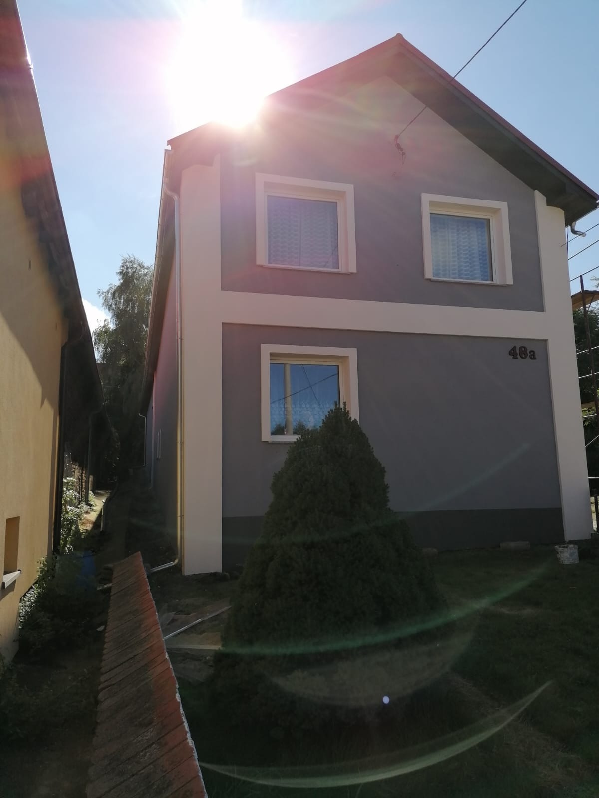 Dwukondygnacyjny dom z szarą elewacją i kremowymi obramowaniami okien oraz narożników, numer domu 48a, widok z boku w słoneczny dzień.