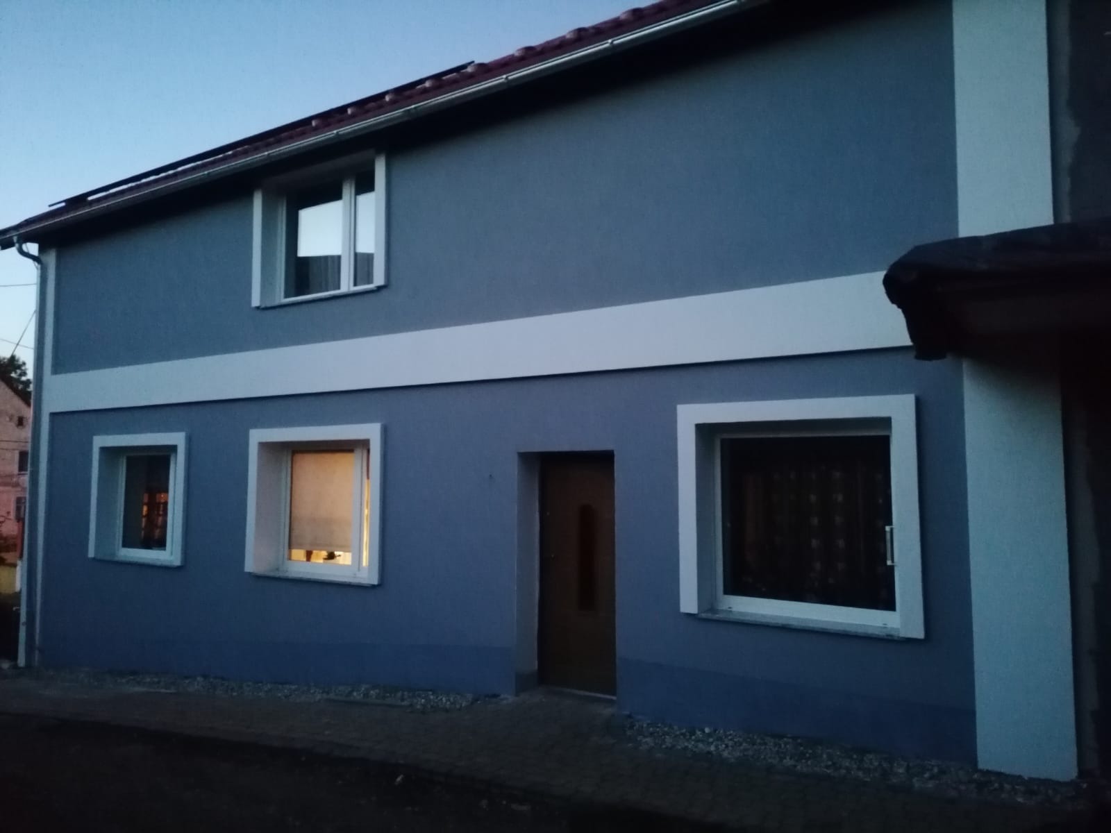 Dwukondygnacyjny dom z niebieską elewacją i białymi obramowaniami okien oraz poziomym pasem, widok z zewnątrz o zmierzchu.