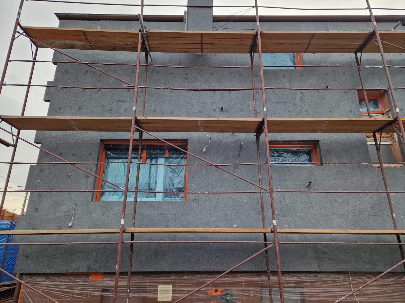 Elewacja budynku w trakcie ocieplania, widoczne rusztowania, szare płyty izolacyjne i zabezpieczone folią okna.