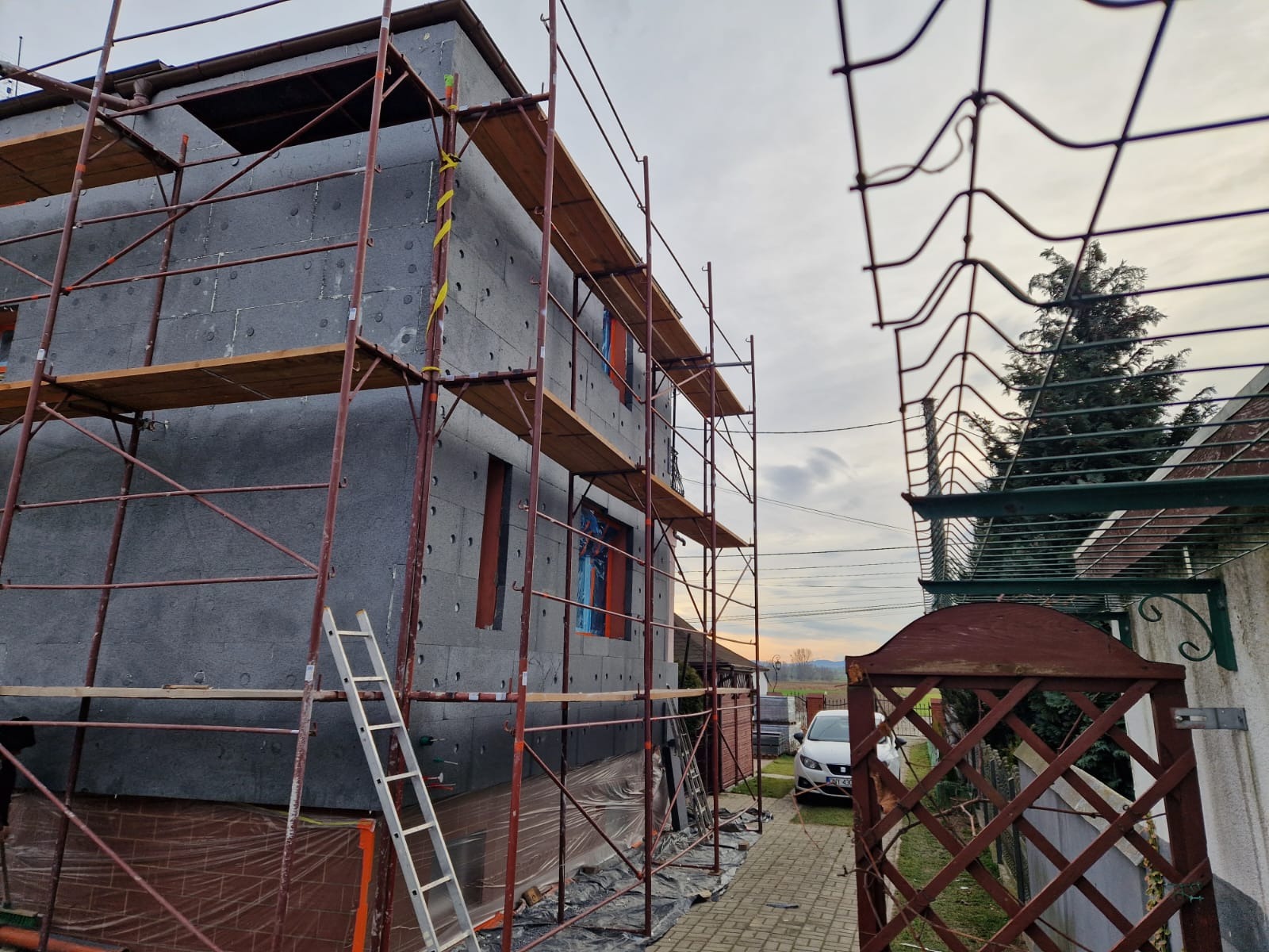 Dom w trakcie ocieplania, widoczne rusztowania, szary styropian na elewacji, drabina oparta o ścianę, brama wejściowa w tle.