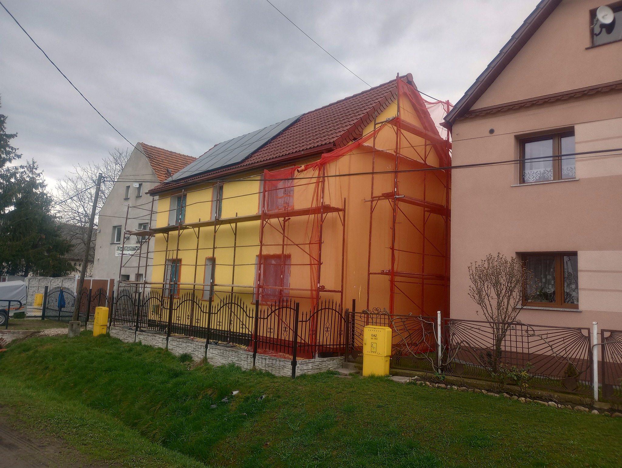 Dom jednorodzinny w trakcie prac elewacyjnych, widoczne rusztowania i siatka ochronna, część elewacji w kolorze żółtym, część w pomarańczowym, na dachu panele fotowoltaiczne.