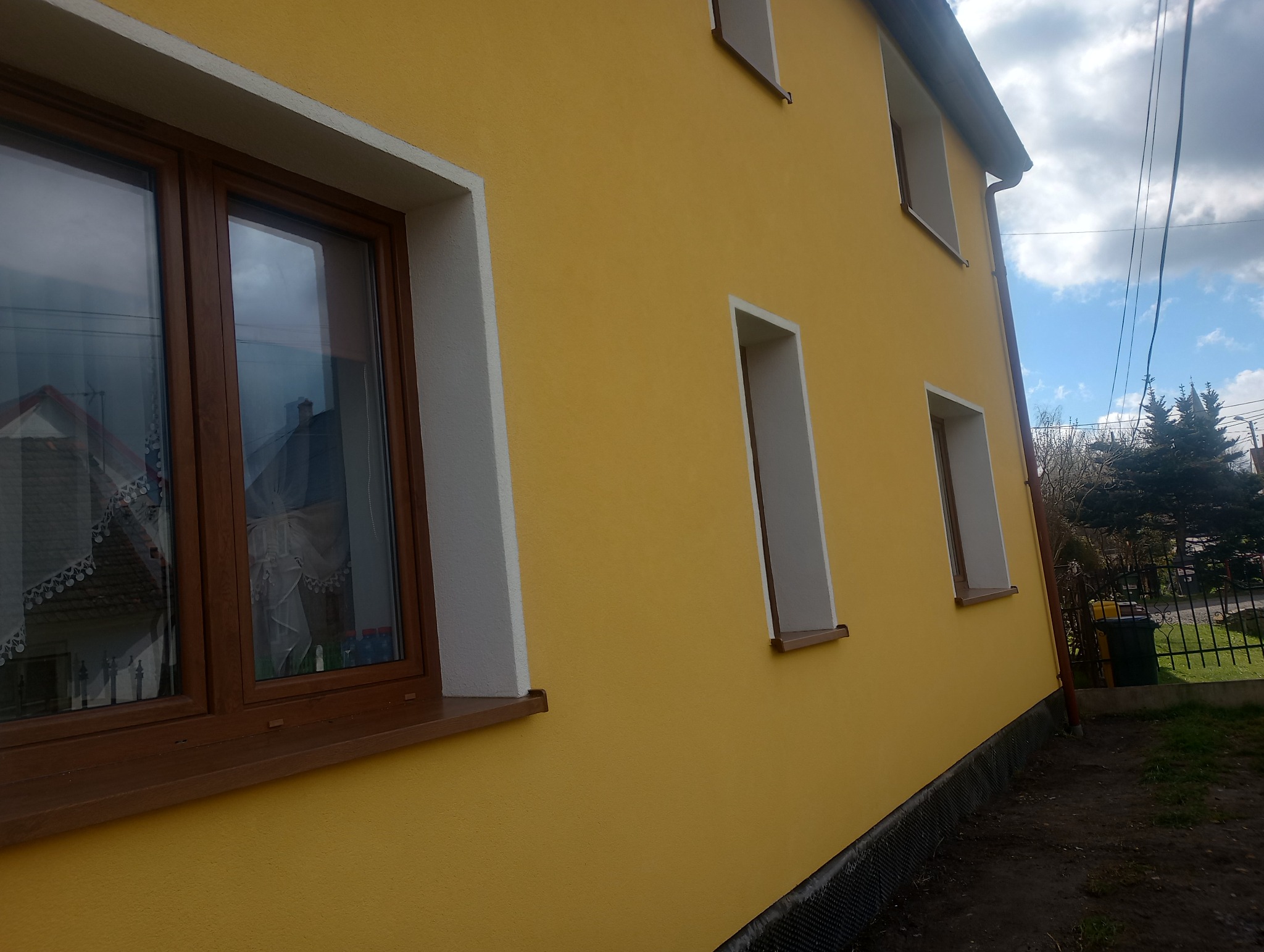 Żółta elewacja domu z brązowymi oknami, widok z boku, z widoczną siatką elewacyjną u dołu ściany.