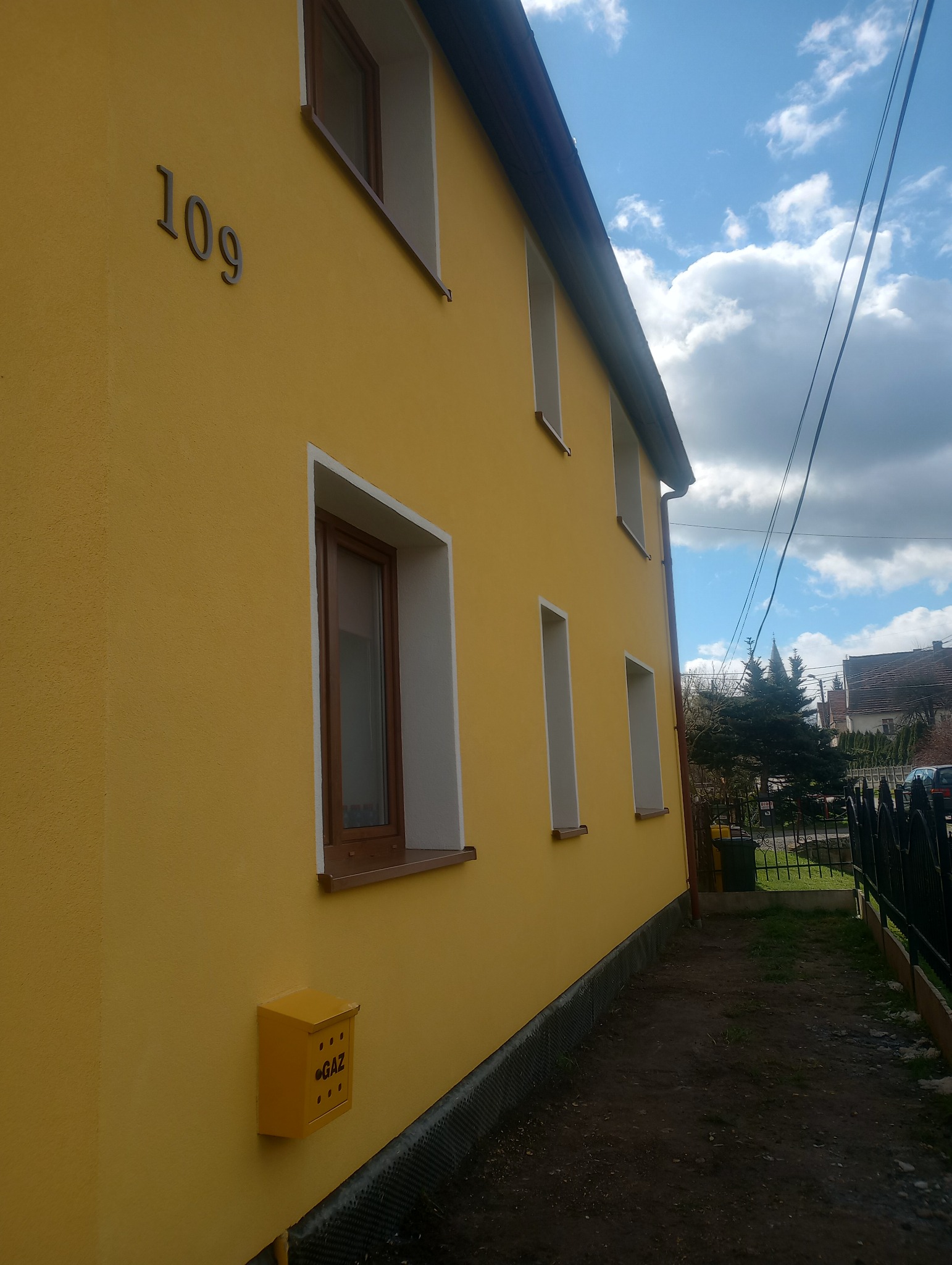 Żółta elewacja domu jednorodzinnego z numerem 109, brązowymi ramami okiennymi i żółtą skrzynką gazową na tle błękitnego nieba z chmurami.