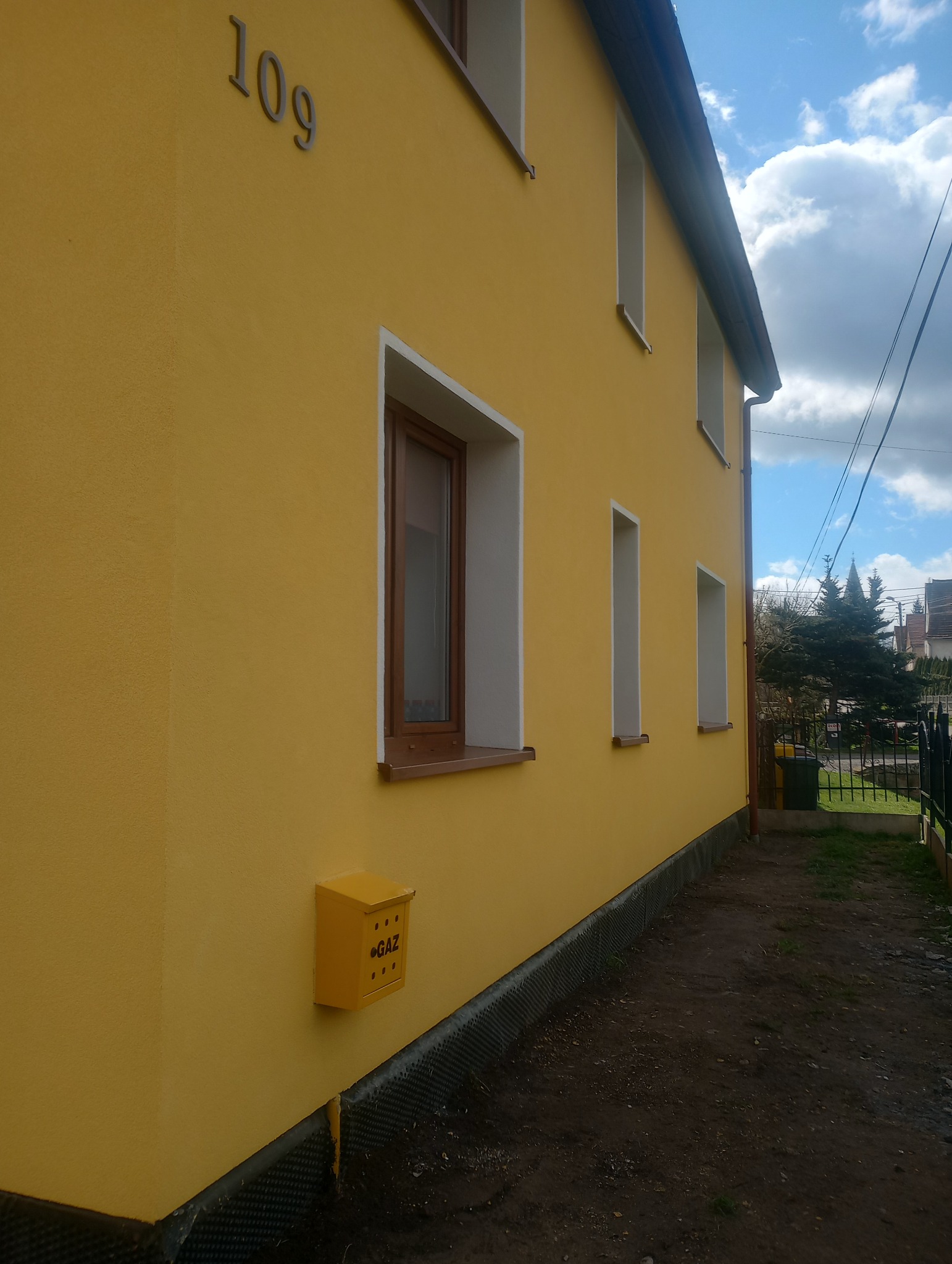 Żółta elewacja dwukondygnacyjnego domu z numerem 109, brązowymi oknami w białych ramach i żółtą skrzynką gazową, widok z boku.