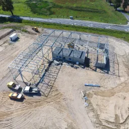 Widok z lotu ptaka na plac budowy hali magazynowej z częścią biurową w Iławie, ukazujący stalową konstrukcję szkieletową budynku w trakcie montażu, stojące obok budynki biurowe, trzy samochody...