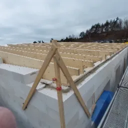 Wznoszenie ścian domu z białych bloczków, widoczne drewniane elementy konstrukcji dachu i rusztowania w tle, pochmurne niebo.