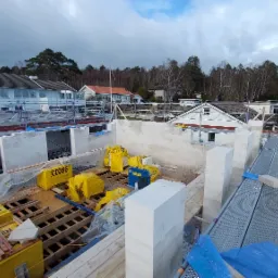 Wznoszenie ścian domu z bloczków Ytong, widoczne rusztowania, narzędzia i materiały budowlane, w tle zabudowa jednorodzinna.