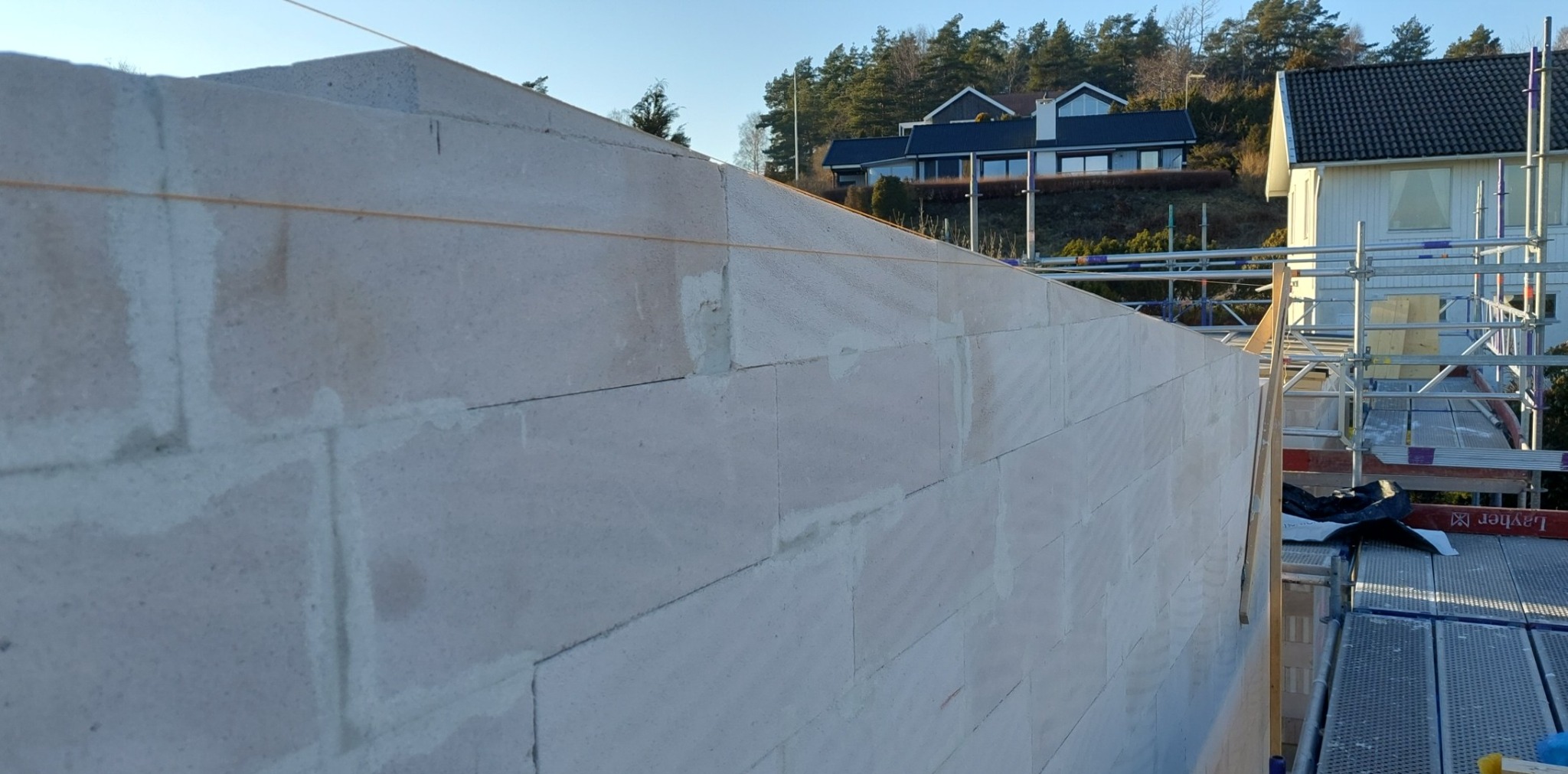 Wznoszona ściana z jasnych bloczków, na tle domów jednorodzinnych i rusztowania.