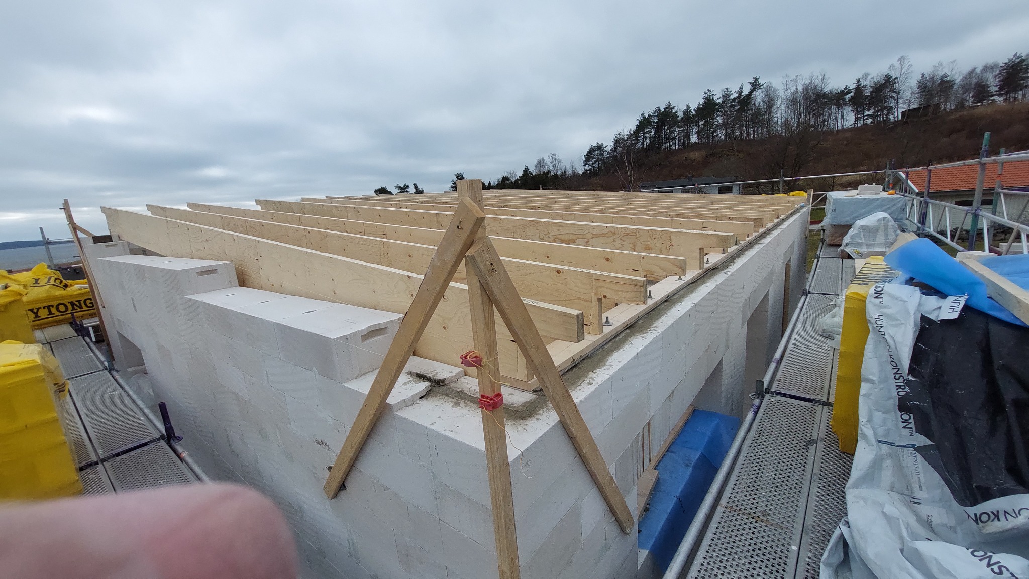 Wznoszenie ścian domu z białych bloczków, widoczne drewniane elementy konstrukcji dachu i rusztowania w tle, pochmurne niebo.