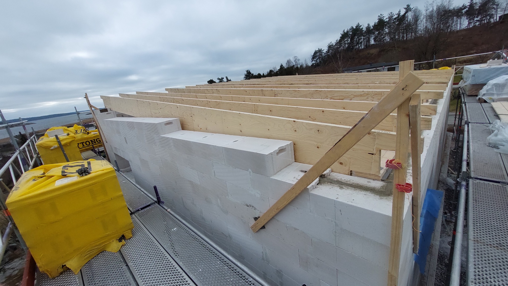 Wznoszenie ścian z białych bloczków oraz montaż drewnianej konstrukcji dachu na placu budowy przy brzegu jeziora, widoczne rusztowania i worki z materiałami budowlanymi.