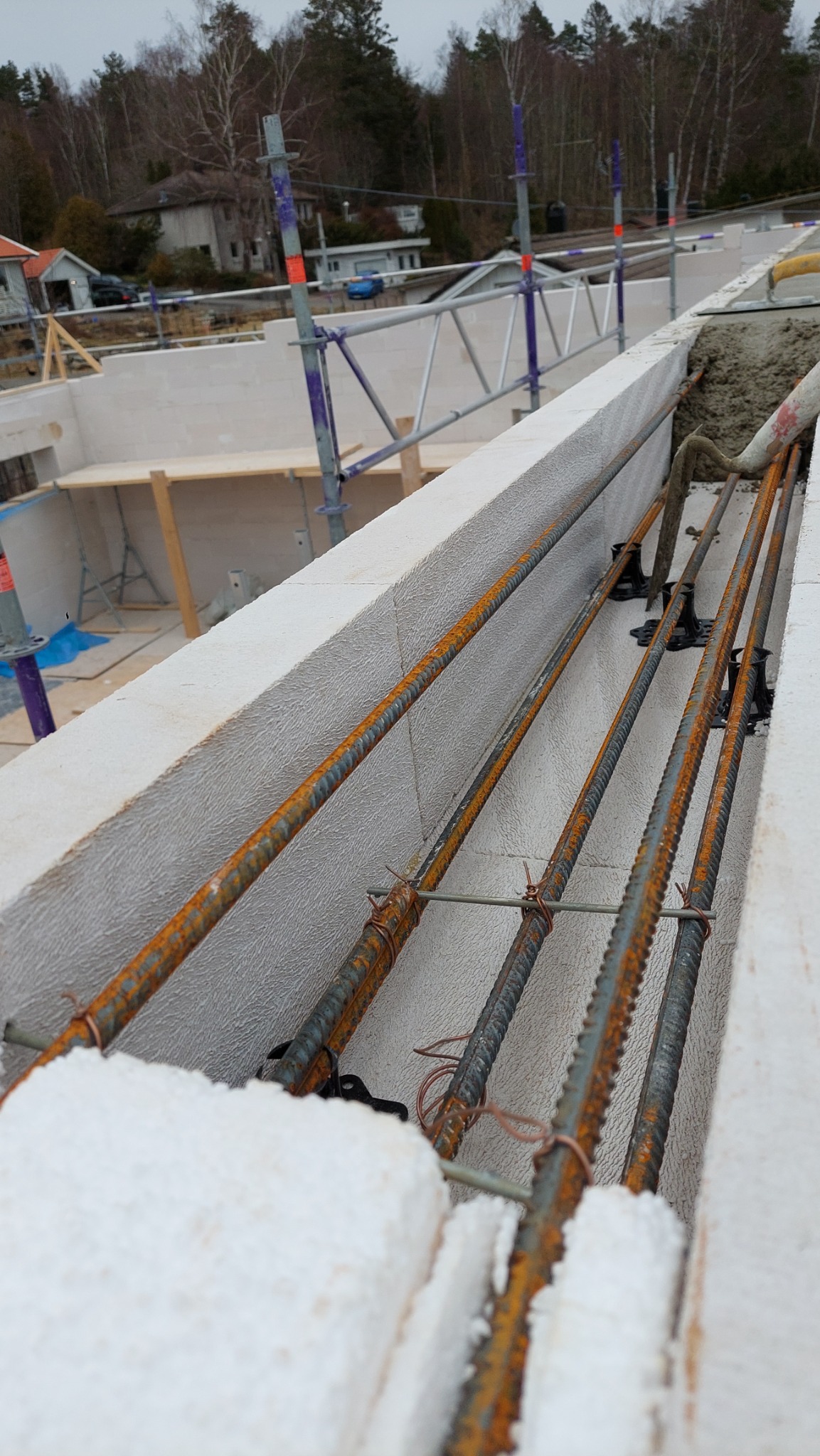 Zbrojenie pionowe i poziome w murze z betonu komórkowego, widoczne elementy rusztowania i otoczenie budowy.