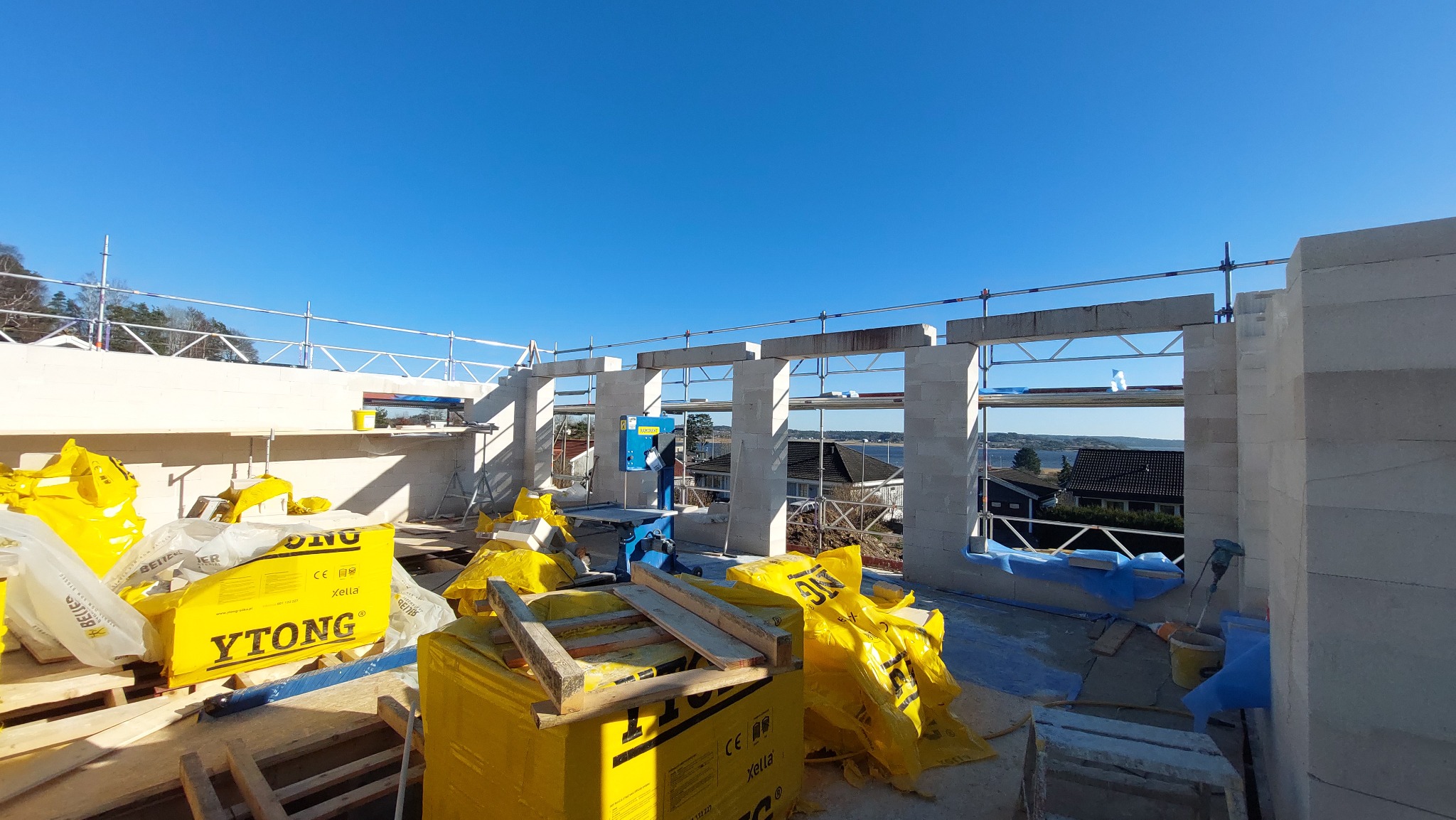 Wznoszenie ścian domu z bloczków Ytong, widoczne narzędzia i materiały budowlane, w tle krajobraz z jeziorem i zabudowaniami.