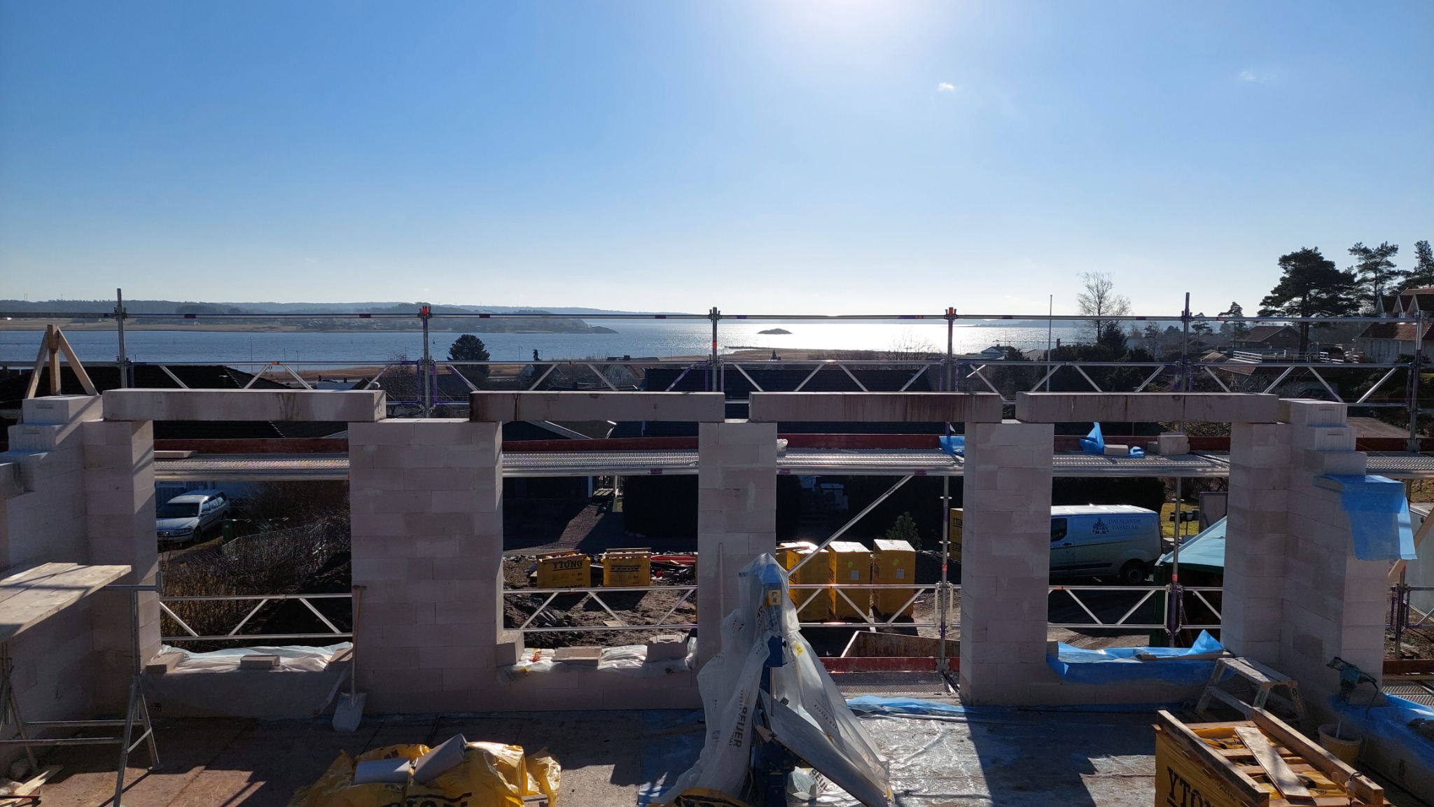 Wznoszenie ścian z bloczków na tle malowniczego krajobrazu z wodą i wyspami, widoczne rusztowania i materiały budowlane.