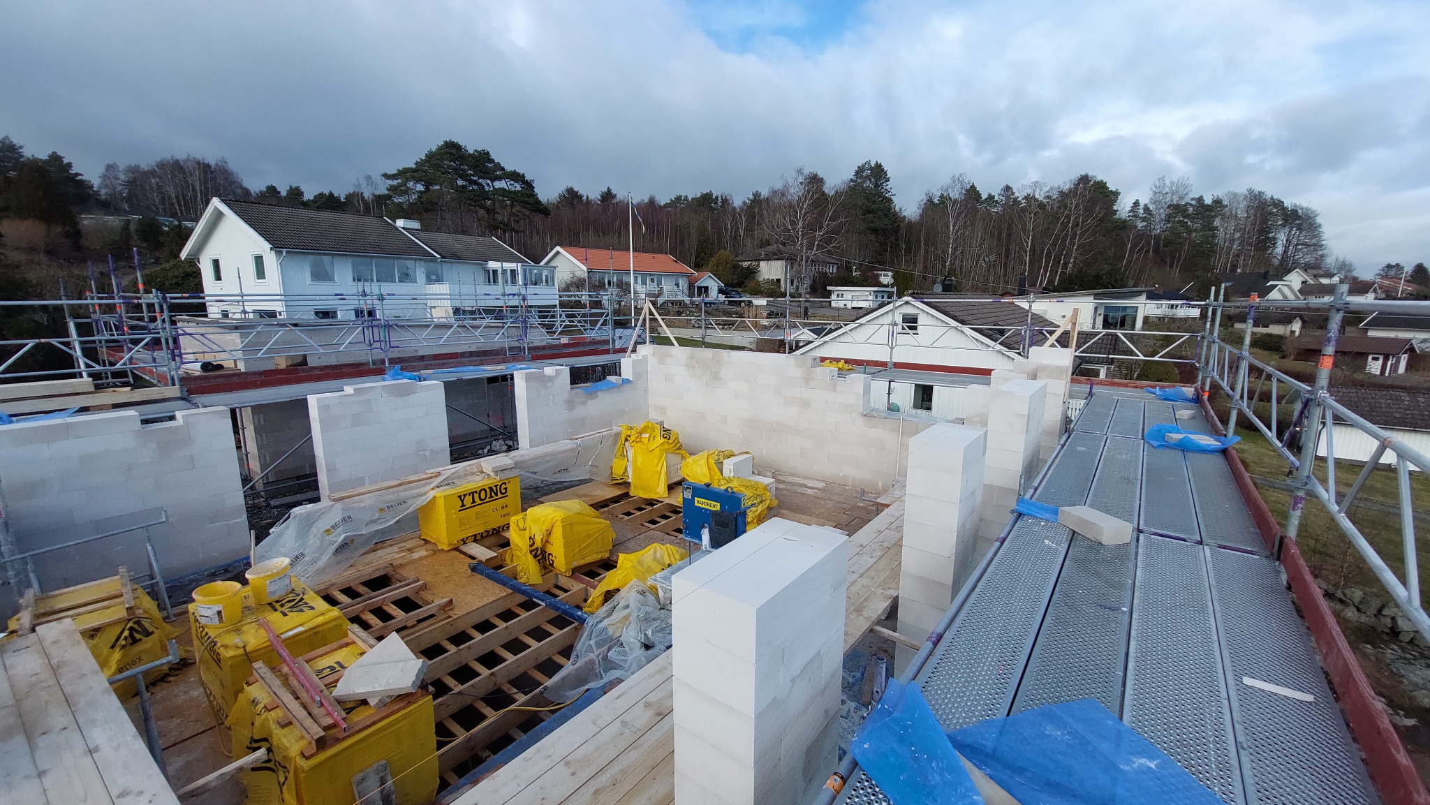 Wznoszenie ścian domu z bloczków Ytong, widoczne rusztowania, narzędzia i materiały budowlane, w tle zabudowa jednorodzinna.