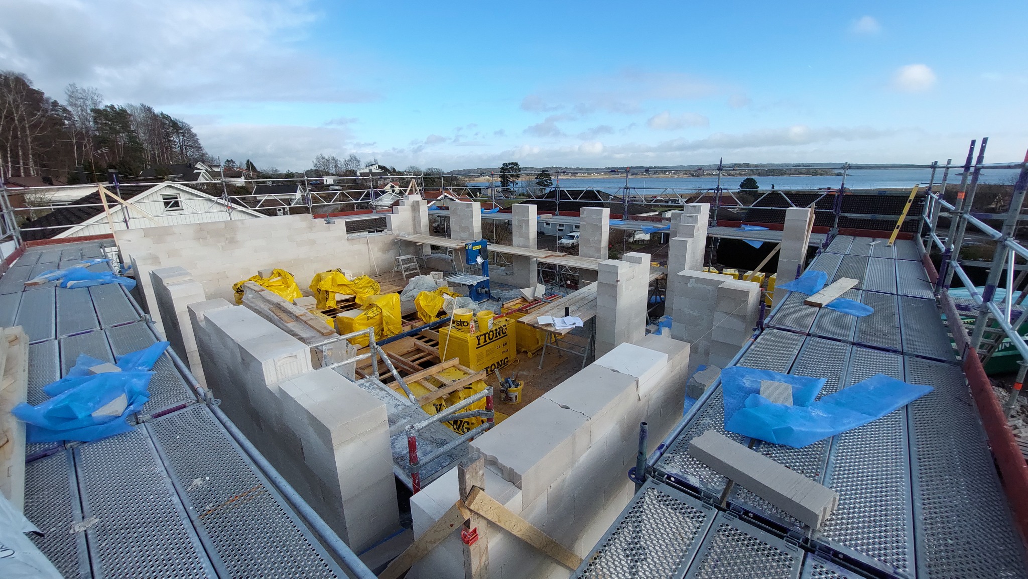 Wznoszenie ścian budynku z bloczków Ytong na tle krajobrazu z jeziorem, widok z rusztowania, materiały budowlane zabezpieczone folią