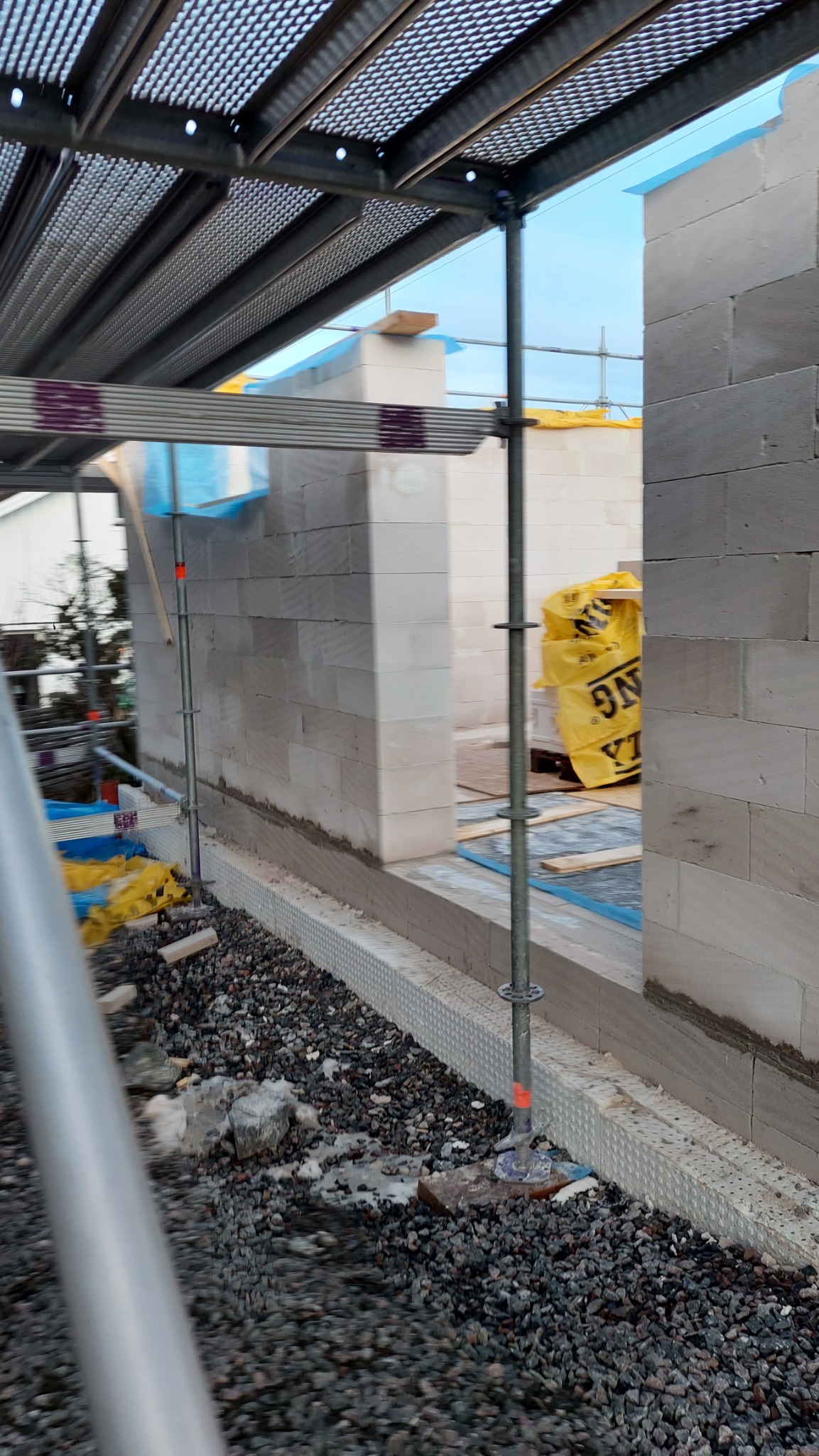 Wznoszenie ścian z bloczków betonowych na fundamencie z izolacją, widoczne rusztowanie i materiały budowlane.