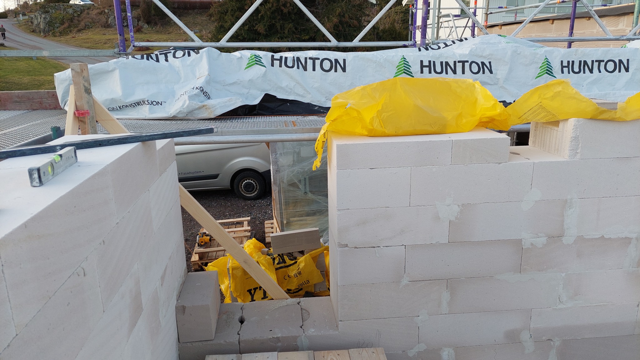 Wznoszenie ściany z białych bloczków, widoczne worki ochronne z materiałami budowlanymi, poziomica i fragment rusztowania.