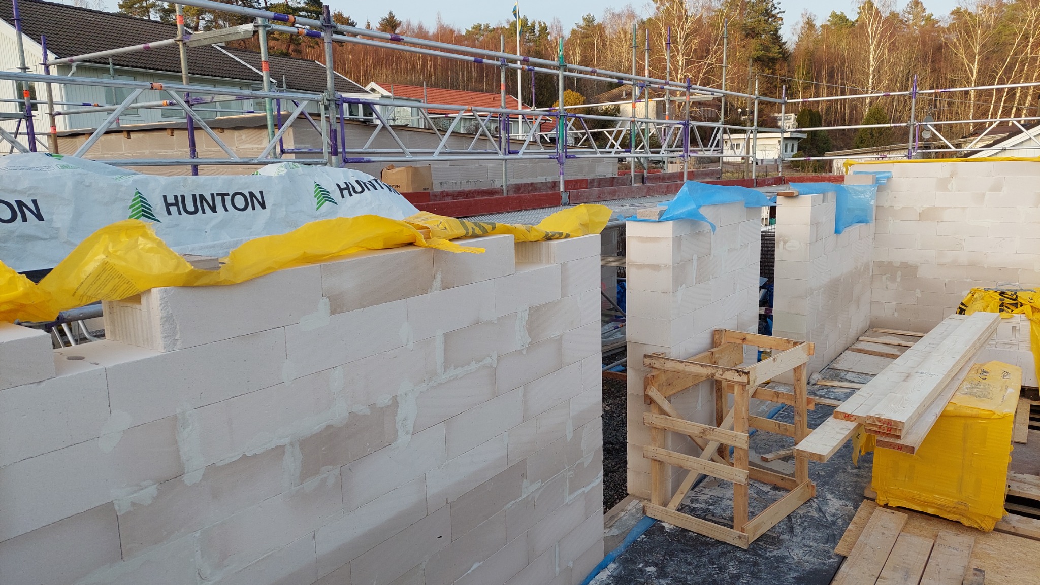 Wznoszone ściany z białych bloczków, z widocznymi spoinami, zabezpieczone folią i rusztowaniami, na tle drzew i zabudowań.