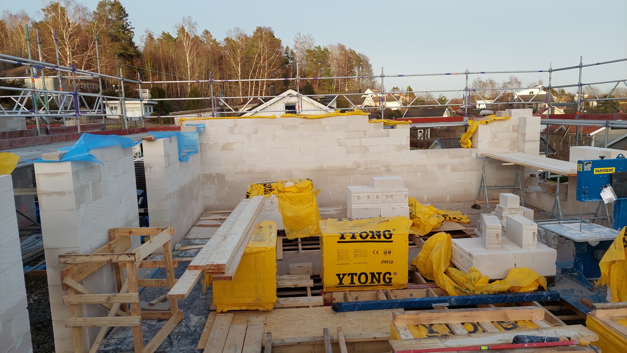 Wznoszenie ścian z bloczków Ytong na budowie domu jednorodzinnego, widoczne rusztowania, materiały budowlane zabezpieczone folią, piła do cięcia bloczków.