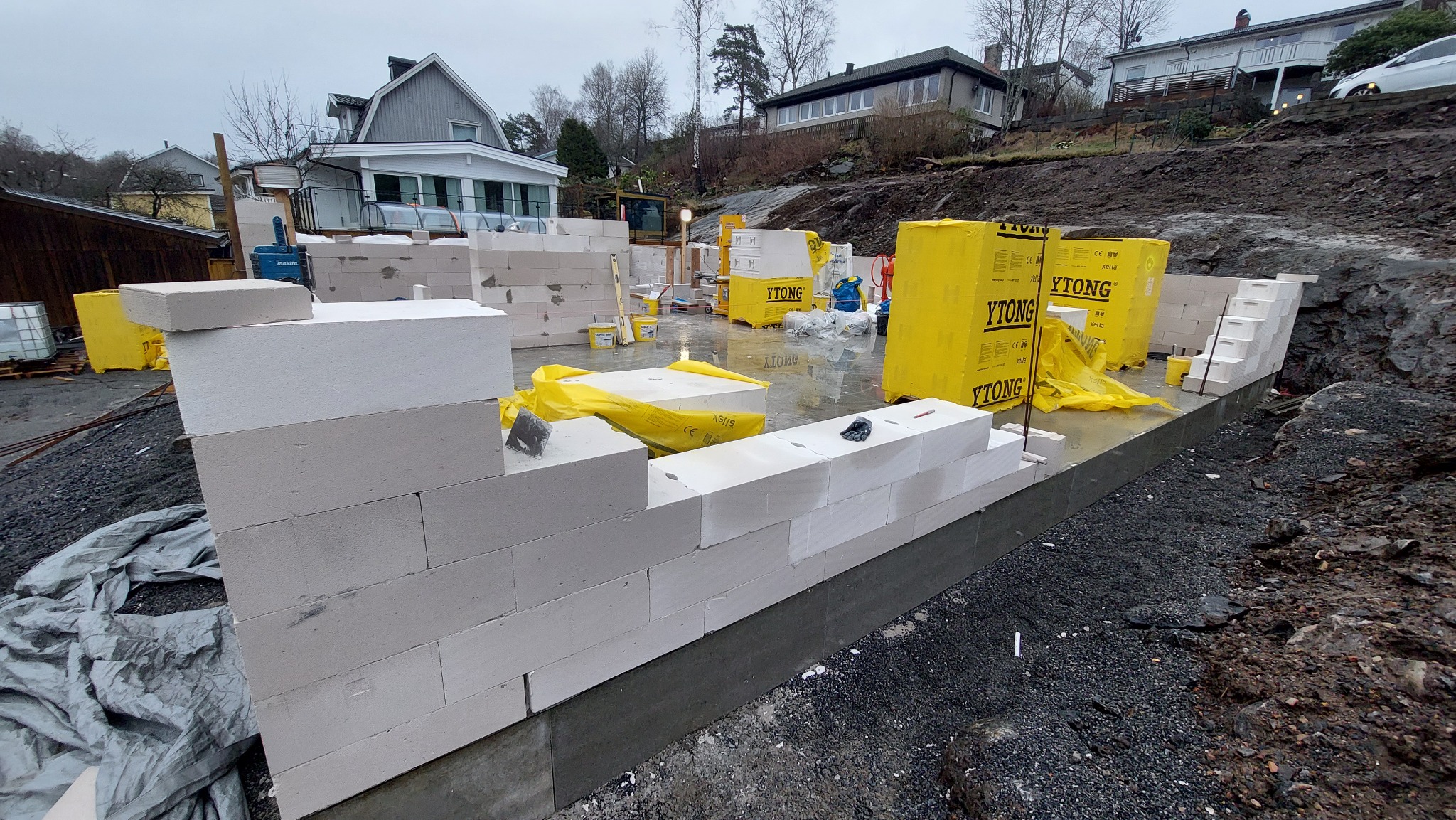 Wznoszenie ścian z białych bloczków Ytong na fundamencie, widoczne palety z bloczkami w żółtej folii, teren budowy z domami w tle.