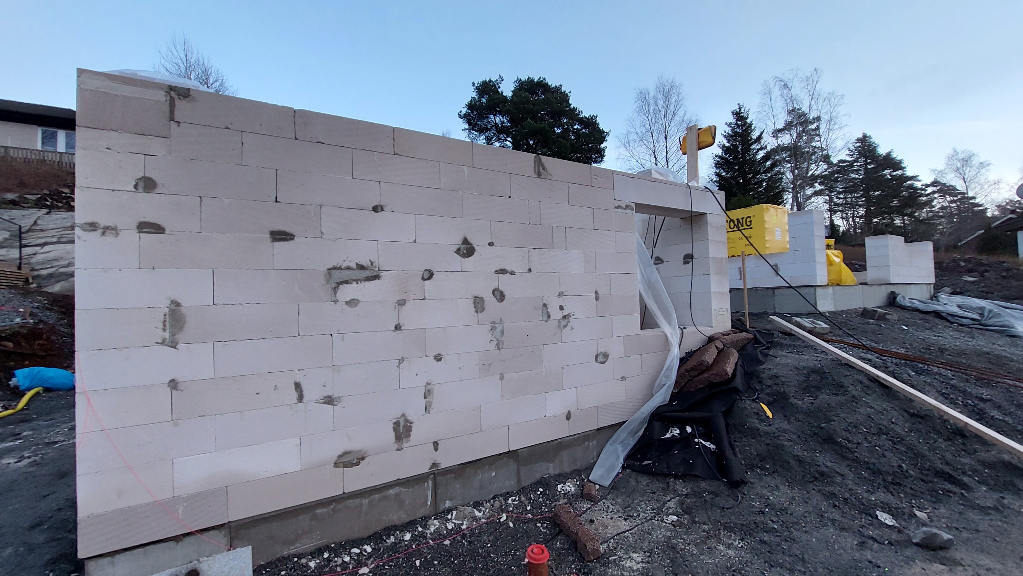 Wznoszenie ściany z betonu komórkowego na fundamencie, widoczne pakiety bloczków oraz teren budowy w tle.