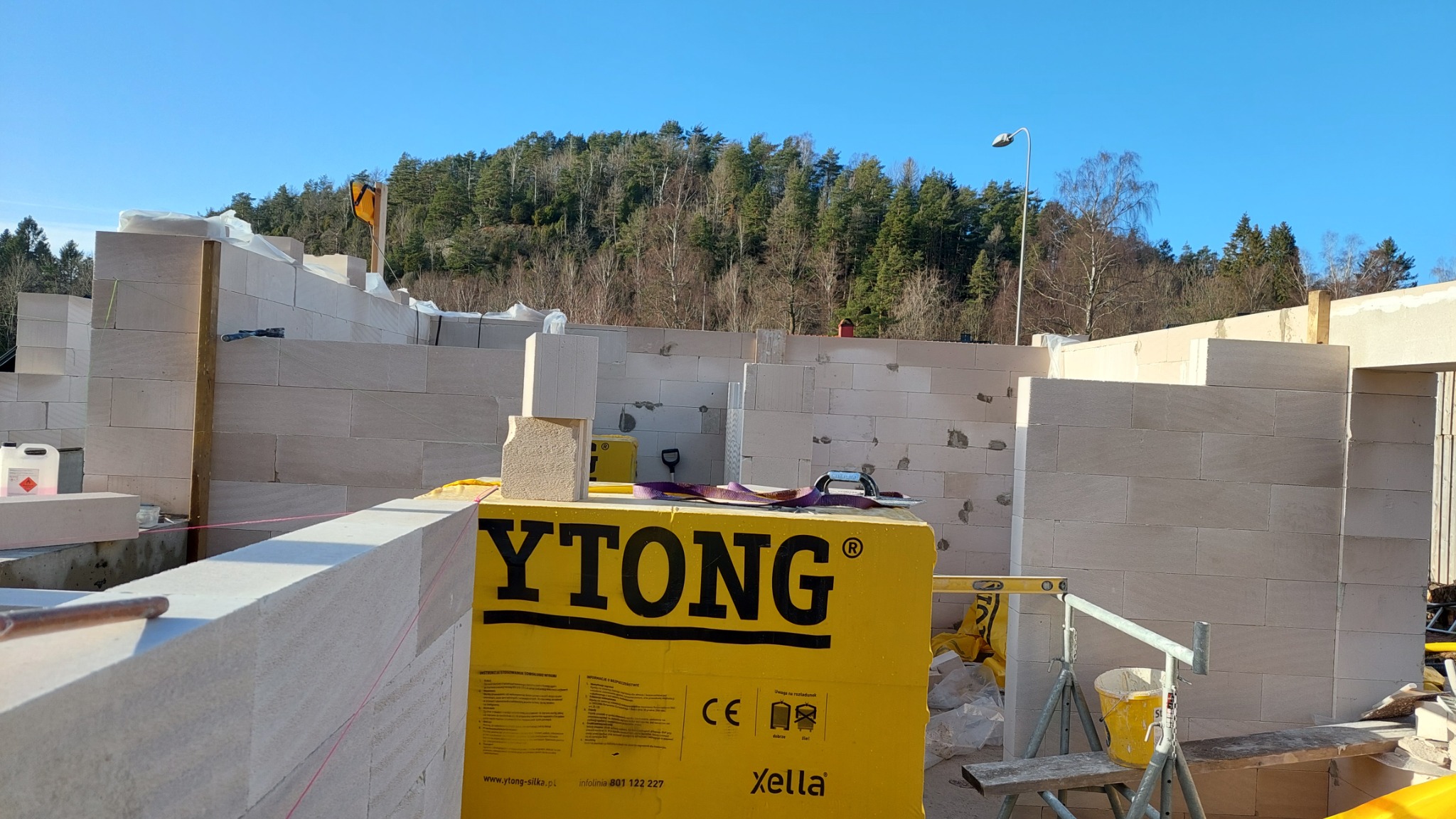 Wznoszenie ścian domu z bloczków Ytong, widoczne narzędzia murarskie, poziomica, las w tle.