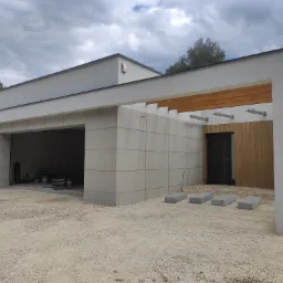 Nowoczesny budynek garażowy z elewacją z płyt betonowych, drewnianymi elementami dekoracyjnymi nad wejściem i żwirowym podłożem, widok na otwarty garaż z narzędziami w środku.