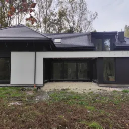 Nowoczesny dom jednorodzinny z czarną elewacją i antracytowym dachem z dachówki ceramicznej, widoczne duże okna i taras przygotowany do wykończenia, otoczenie w trakcie zagospodarowywania.