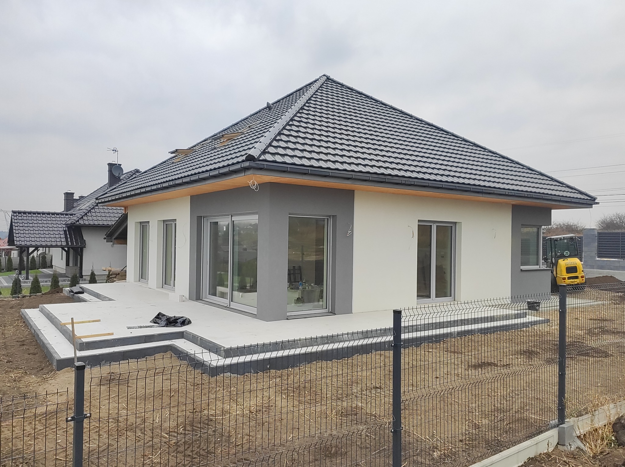 Nowoczesny dom jednorodzinny z szarą dachówką i biało-szarą elewacją, widoczny częściowo ukończony taras z betonowych płyt oraz mała koparka na posesji.