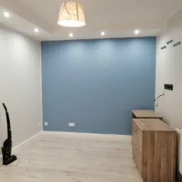 Pomieszczenie po malowaniu ścian na biało i niebiesko, widoczne nowe listwy przypodłogowe, lampa sufitowa i odkurzacz pionowy.