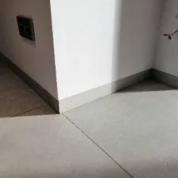 Szare płytki podłogowe z fugami, listwa przypodłogowa w kolorze płytek, białe ściany z szarą listwą przy podłodze, widoczny fragment gniazdka elektrycznego.