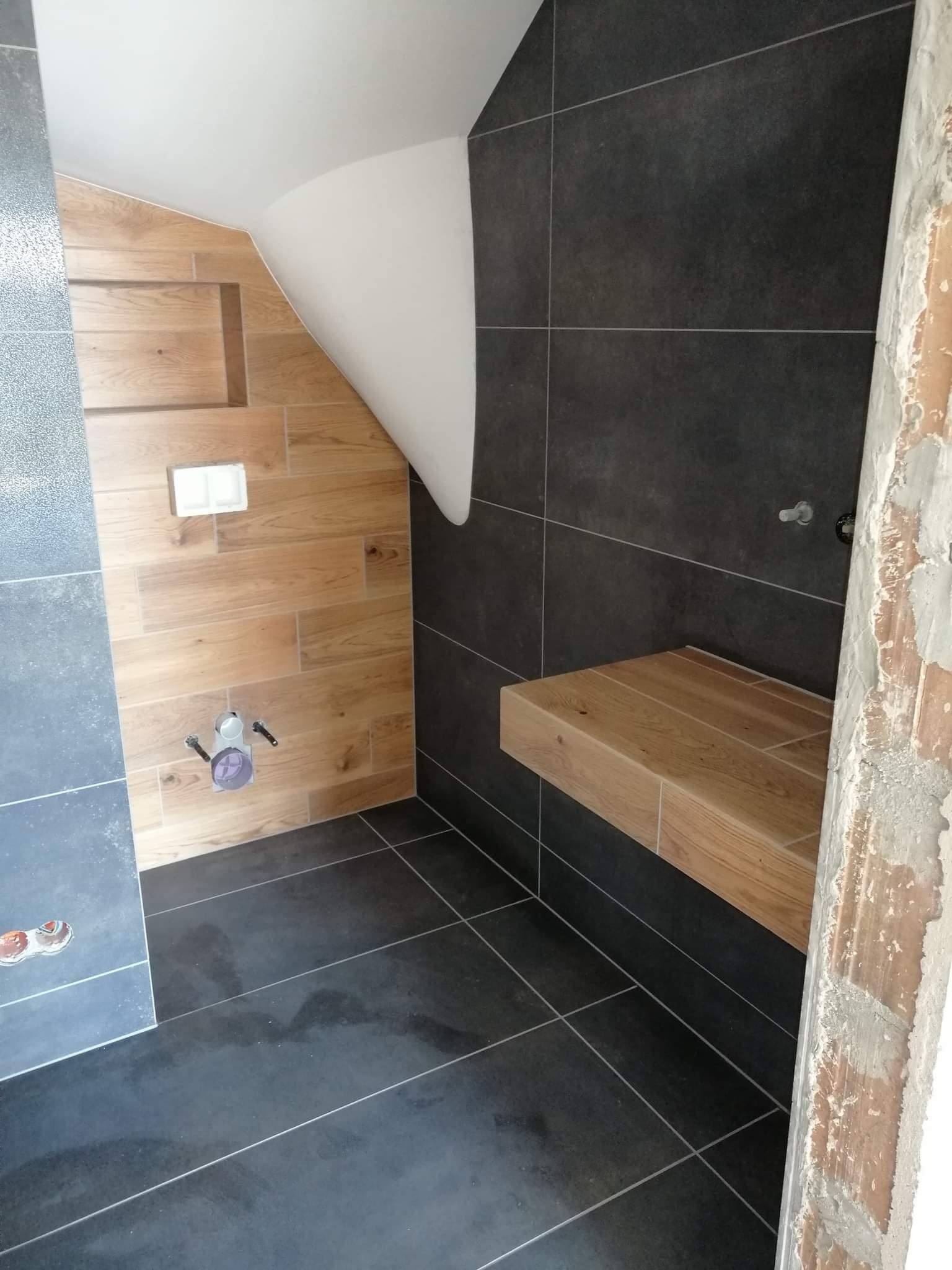 Łazienka w trakcie wykańczania: ciemne płytki podłogowe i ścienne kontrastują z drewnopodobnymi płytkami na ścianie ze skosem i wnęką, widoczny fragment niezabezpieczonego otworu drzwiowego.