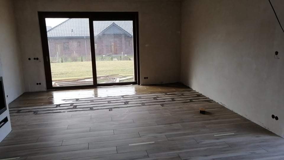 Wnętrze w trakcie układania podłogi z płytek imitujących drewno; widoczne fragmenty kleju i listwy dystansowe.