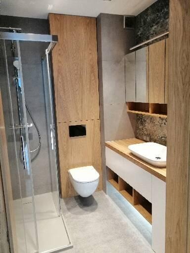 Nowoczesna łazienka z kabiną prysznicową ze srebrnymi profilami, podwieszaną toaletą, szafką z umywalką nablatową i drewnianymi akcentami.