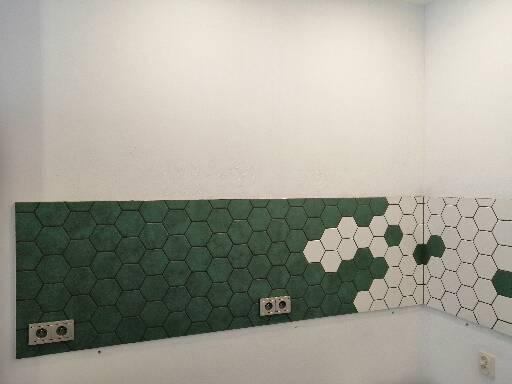 Fragment ściany wyłożonej heksagonalnymi płytkami w odcieniach zieleni i bieli, z widocznymi gniazdkami elektrycznymi, tworzący mozaikowy wzór.