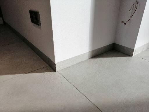 Szare płytki podłogowe z fugami, listwa przypodłogowa w kolorze płytek, białe ściany z szarą listwą przy podłodze, widoczny fragment gniazdka elektrycznego.