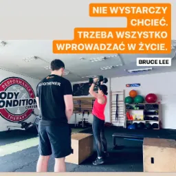 Kobieta w różowej koszulce i czarnych legginsach podnosi hantle pod okiem trenera w czarnej koszulce z napisem 'FIZJOTRENER', w tle widoczne wyposażenie siłowni i cytat Bruce'a Lee o motywacji.