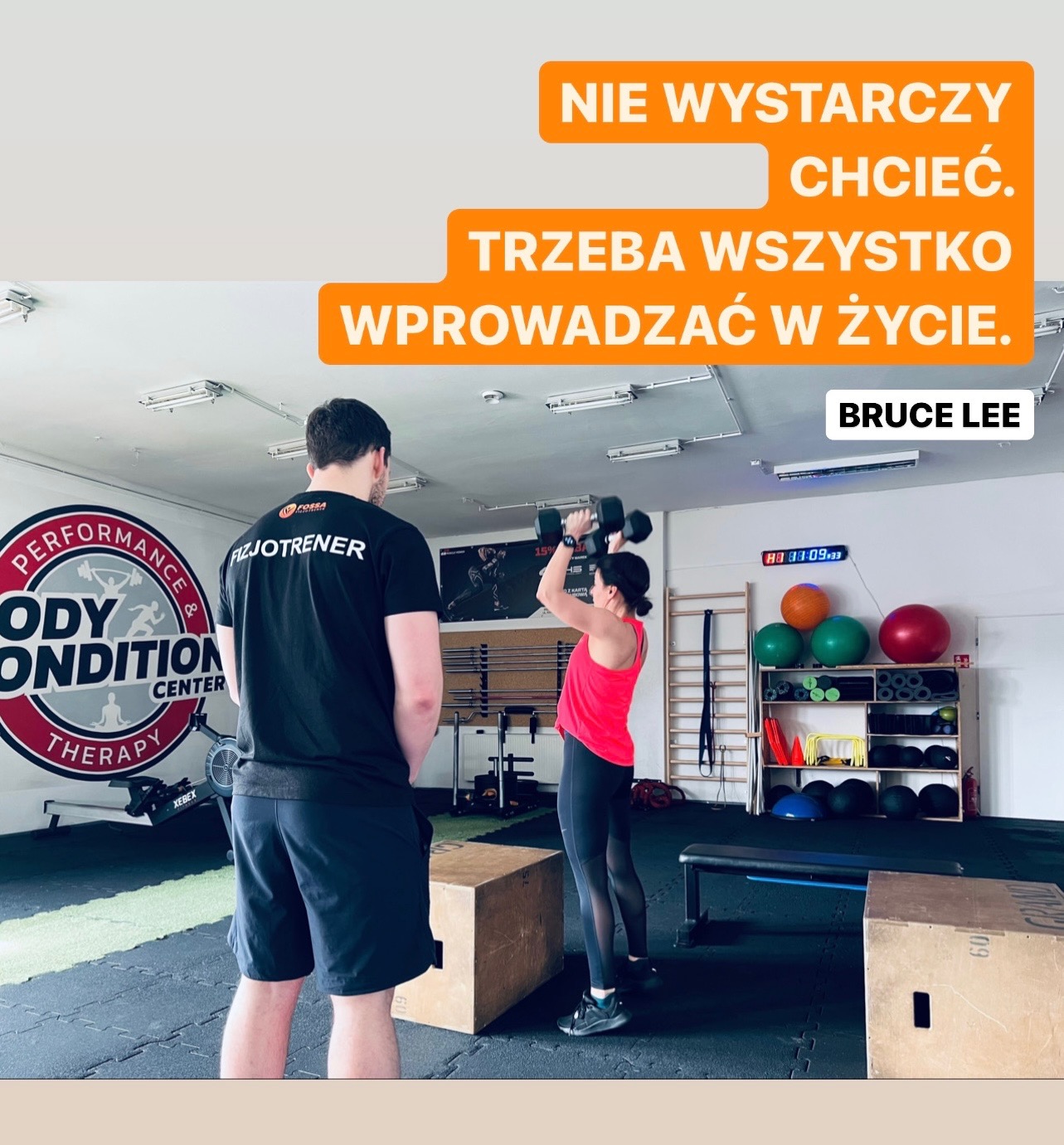 Kobieta w różowej koszulce i czarnych legginsach podnosi hantle pod okiem trenera w czarnej koszulce z napisem 'FIZJOTRENER', w tle widoczne wyposażenie siłowni i cytat Bruce'a Lee o motywacji.