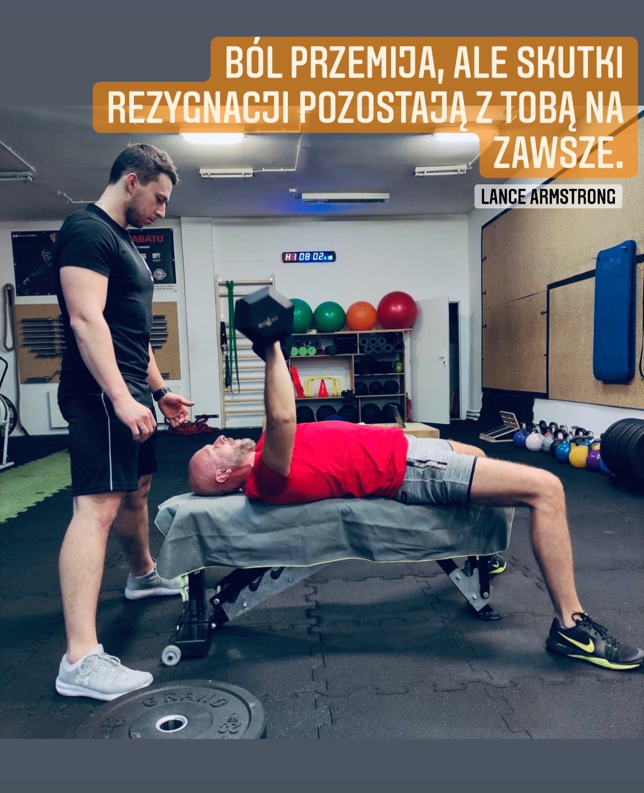 Mężczyzna leży na ławeczce, podnosi ciężarek jedną ręką, drugi mężczyzna stoi obok i obserwuje, w tle sala ćwiczeń z piłkami i sprzętem.