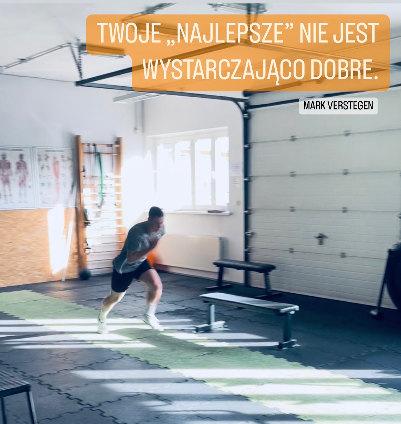 Mężczyzna w szarym t-shircie i czarnych spodenkach biegnie po zielonym pasie na czarnej podłodze w przestronnym pomieszczeniu z białymi ścianami, oknem i podnoszoną bramą garażową; widoczne...